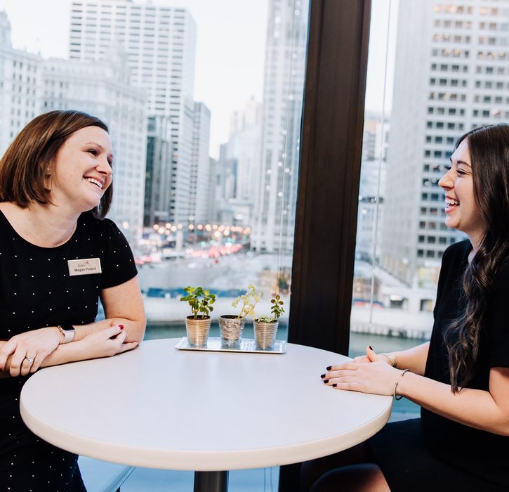 Two women talking in front of a large window with skyscrapers in the background